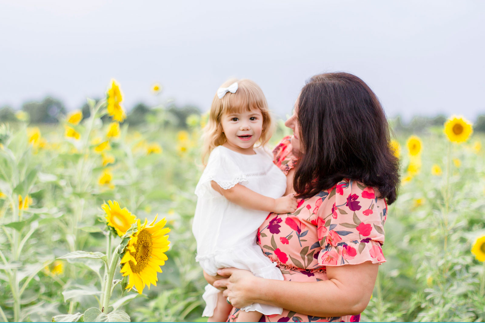 mom looking at her daughter as the little girl looks at the camera by the sunflowers at the sunflower field in Belcher, LA