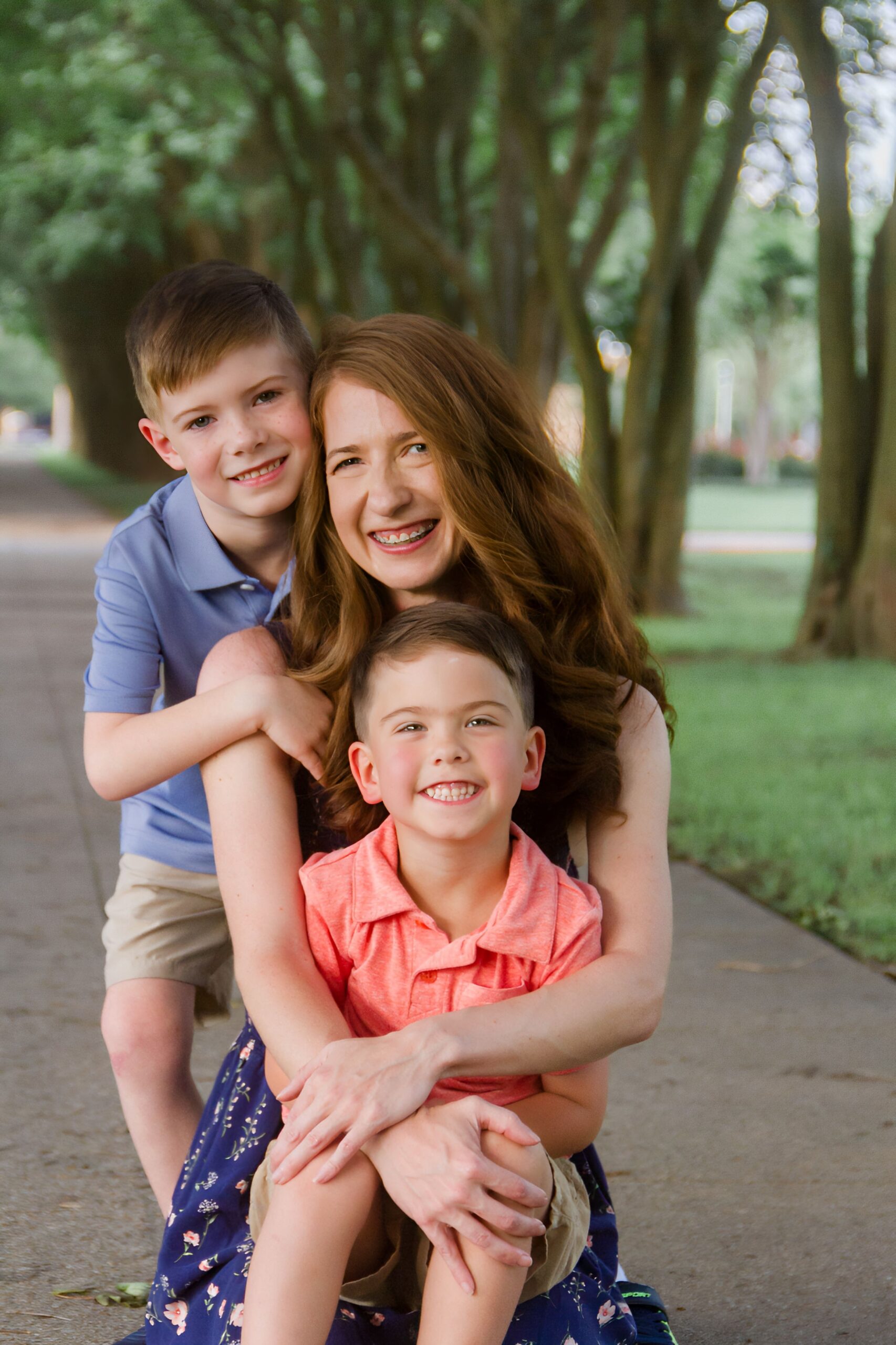 mom holding one son on her knees while her other son is hugging her from behind on a pathway between trees outdoor for a family session in Shreveport LA