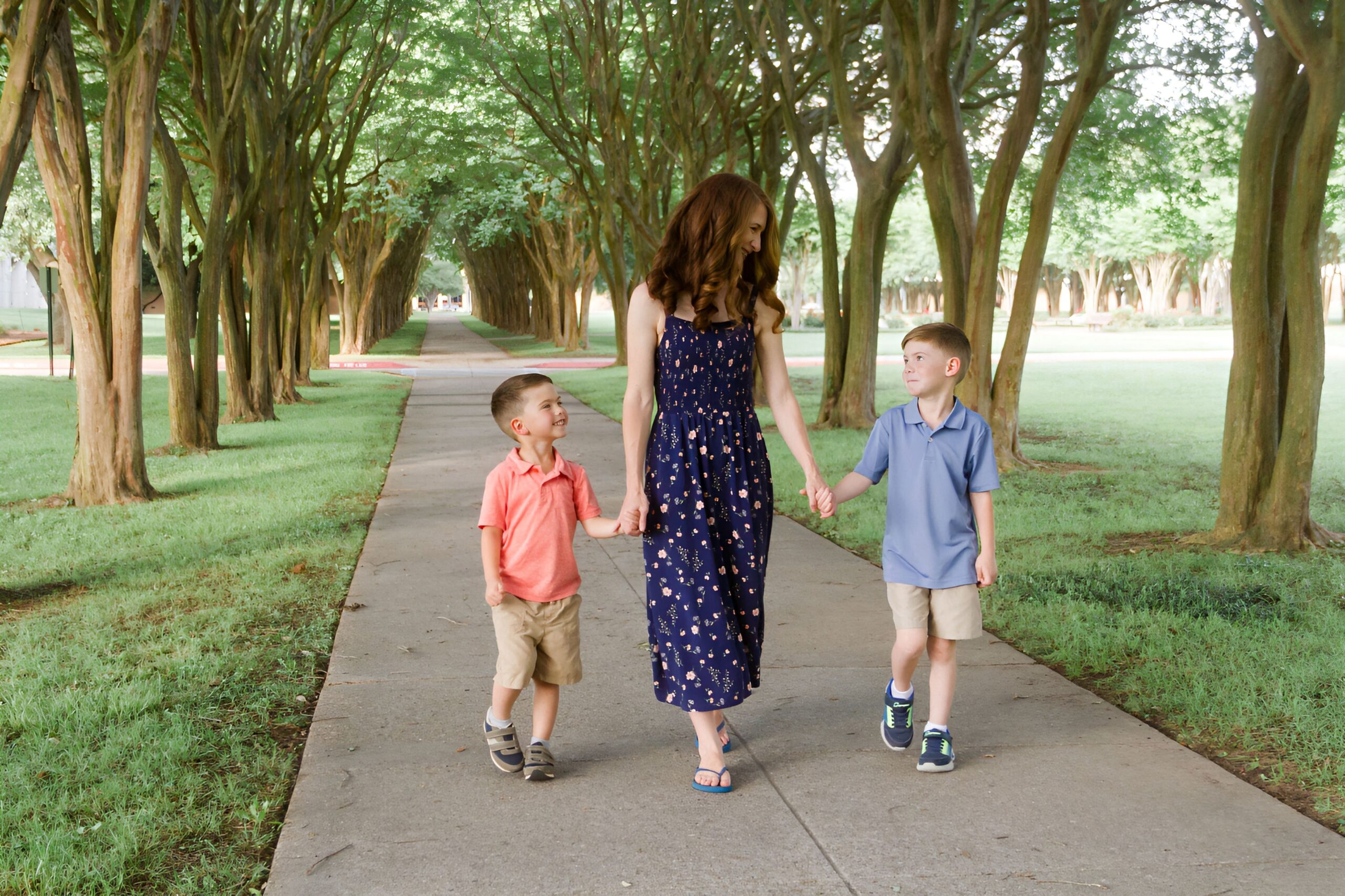 mom holding her two sons hands while walking down on a pathway between trees outdoor for a family session in Shreveport LA