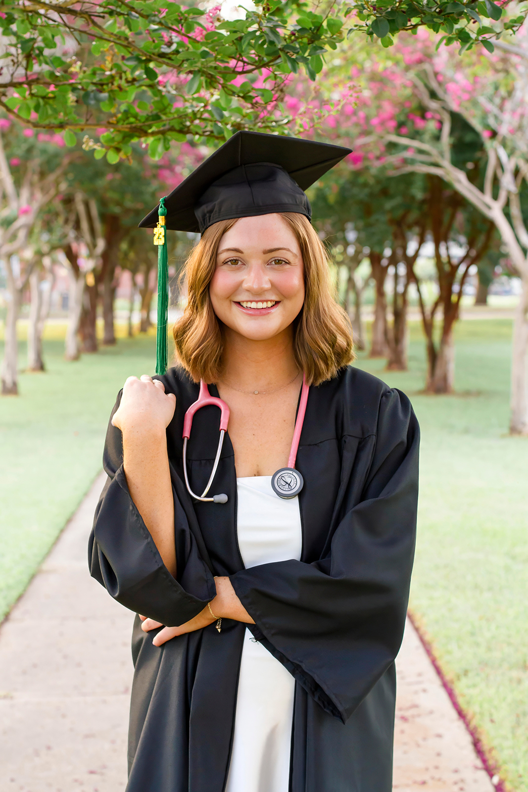 college grad med senior standing near pink trees wearing black cap and gown with pink stethoscope around her neck on LSUS campus Shreveport LA